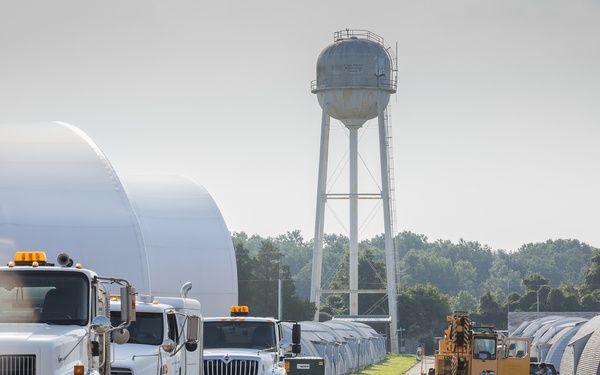 The image shows an industrial or logistics facility with multiple white semi-trucks (cab-over and conventional configurations) lined up along a paved road. The facility features large white dome-shaped structures on the left and Quonset hut-style metal buildings on the right, with a water tower visible in the background. Workers and heavy equipment, including a yellow crane or lift vehicle, are active in the area.