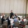 2021 Army Strong Bonds for Kentucky National Guard A man in a black polo shirt stands and appears to be speaking or facilitating a session in a banquet-style room. Multiple participants, appearing to be a mix of adults and younger individuals, are seated at round tables covered with white tablecloths. The setting resembles a workshop, training session, or educational event in a conference or event hall.