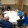 2021 Army Strong Bonds for Kentucky National Guard A group of adults and children are gathered around a round table in what appears to be a conference or meeting room. They are engaged in a collaborative activity involving writing on large sheets of white paper spread across the table. The group includes families with young children, and the atmosphere appears casual and interactive.
