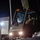 U.S. Navy Seabees assigned to NMCB-5 support the Defense Logistics Agency A person wearing camouflage military-style clothing and a cap is seated in the cab of a large military or industrial vehicle at night. The vehicle's headlights are illuminated, and a crane or boom arm is visible extending upward from the vehicle. The scene appears to be at a military installation or industrial facility.