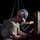 U.S. Navy Seabees assigned to NMCB-5 support the Defense Logistics Agency A military service member, likely U.S. Navy based on the digital camouflage (NWU Type I) trousers, works at night performing a hands-on task near heavy industrial equipment. The individual is wearing full personal protective equipment including a hard hat with a unit insignia sticker, yellow safety glasses pushed up on the helmet, a black face mask, and gloves. The scene appears to be a nighttime working party or maintenance operation on a pier, dock, or vessel.