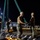 US Navy Seabees assigned to NMCB-5 support the Defense Logistics Agency A group of military personnel, appearing to be U.S. Navy sailors based on their camouflage uniforms, are conducting a nighttime logistics or rigging operation at what appears to be a port or naval facility. They are working with heavy lifting straps and shackles, likely securing or moving large cylindrical objects arranged in rows. All personnel are wearing hard hats and face masks, suggesting this is a safety-regulated work environment.