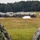 A military field camp is being established on an open grassy area bordered by dense forest. Personnel in camouflage uniforms are actively setting up or arranging a large row of military-style tents across the field. The foreground shows blurred camouflage fabric, likely from tents or gear in the immediate vicinity of the camera.