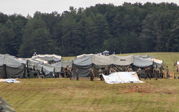 A military field camp is being established on an open grassy area bordered by dense forest. Personnel in camouflage uniforms are actively setting up or arranging a large row of military-style tents across the field. The foreground shows blurred camouflage fabric, likely from tents or gear in the immediate vicinity of the camera.