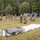 A group of military personnel in camouflage uniforms are assembling a large tent or temporary shelter structure on an open grassy field. The frame of the structure is partially erected while tent canvas material lies on the ground nearby. A row of portable toilets is visible in the background, suggesting a temporary field camp or training exercise setup.