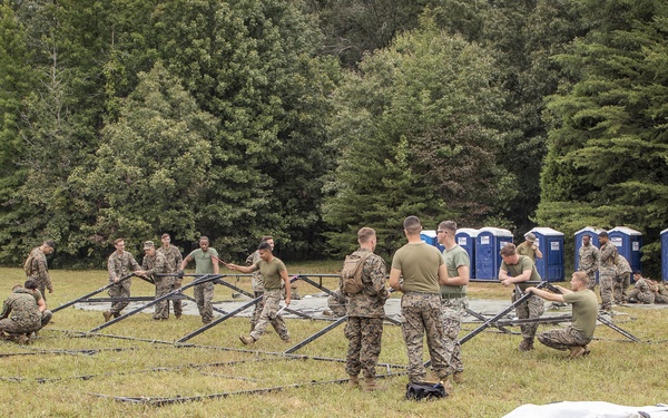 A group of military personnel in camouflage uniforms are assembling a large tent or temporary shelter structure on an open grassy field. The frame of the structure is partially erected while tent canvas material lies on the ground nearby. A row of portable toilets is visible in the background, suggesting a temporary field camp or training exercise setup.