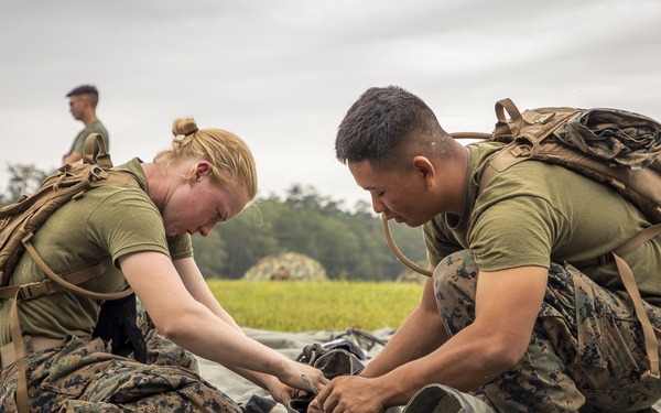 Two military personnel in U.S. Marine Corps MARPAT camouflage uniforms are working together to set up or pack a large tarp or tent on a grassy field. Both are wearing tactical backpacks and are kneeling on the ground, focused on the task. Additional personnel and partially erected shelters are visible in the background.
