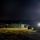 A nighttime military field camp is illuminated by portable light towers and equipment lighting. Several soldiers in military uniforms are gathered near a row of olive-green military tents set up on a grassy field. The scene depicts an active field operation or training exercise with various military supplies and containers visible.