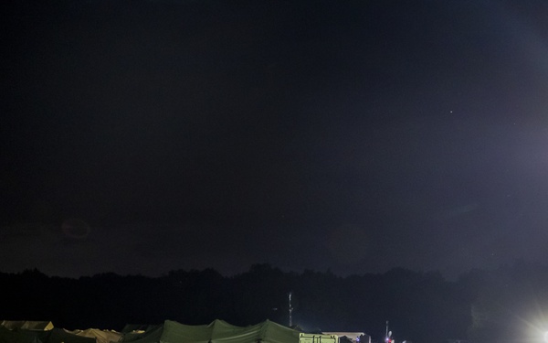 A nighttime military field camp is illuminated by portable light towers and equipment lighting. Several soldiers in military uniforms are gathered near a row of olive-green military tents set up on a grassy field. The scene depicts an active field operation or training exercise with various military supplies and containers visible.