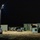 A nighttime military field operation showing personnel working among military equipment and containers illuminated by a portable light tower. Two soldiers in camouflage uniforms are visible near olive-drab military storage containers and HVAC units. The scene appears to be a temporary forward operating base or field logistics area.