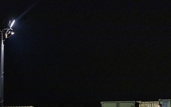 A nighttime military field operation showing personnel working among military equipment and containers illuminated by a portable light tower. Two soldiers in camouflage uniforms are visible near olive-drab military storage containers and HVAC units. The scene appears to be a temporary forward operating base or field logistics area.