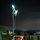 A military service member in camouflage uniform operates a portable light tower at night in what appears to be a forward operating base or field camp. The light tower, powered by a generator unit with visible red indicator lights, illuminates the area with multiple bright floodlights mounted on an extendable mast. Multiple military-style green tents and equipment crates are visible in the background.