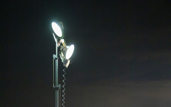 A military service member in camouflage uniform operates a portable light tower at night in what appears to be a forward operating base or field camp. The light tower, powered by a generator unit with visible red indicator lights, illuminates the area with multiple bright floodlights mounted on an extendable mast. Multiple military-style green tents and equipment crates are visible in the background.