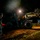 A nighttime military logistics operation showing personnel working alongside a military rough-terrain forklift or loader vehicle. The scene is illuminated primarily by the vehicle's headlights against a dark, wooded background. At least two uniformed soldiers are visible, with supply containers or crates nearby.
