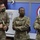 Three U.S. Army soldiers in OCP (Operational Camouflage Pattern) uniforms are engaged in conversation in what appears to be an indoor military facility. All three are wearing face masks, suggesting this was taken during the COVID-19 pandemic period. Name tapes visible on the uniforms read 'GALLUCCI' (left), 'JACKSON' (center), and 'CLARK' (right).