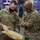 Two U.S. Army soldiers in OCP (Operational Camouflage Pattern) uniforms and black face masks are examining a yellow sealed package, likely an MRE (Meal Ready-to-Eat) or similar military supply item. The soldier on the left is identified by his name tape as 'GLATHAR,' while the soldier on the right wears a Ranger tab and a First Army patch. They appear to be in an office or administrative workspace environment.