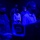 A group of approximately six individuals are gathered in a darkened room illuminated by blue lighting, all wearing black face masks. Some individuals appear to be seated at computer workstations while others stand observing in the background. The setting suggests a control room, operations center, or training facility environment.