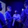 Four individuals are gathered in a dimly lit room with blue ambient lighting, engaged in a discussion. All participants are wearing face masks, suggesting this was taken during the COVID-19 pandemic period. A large display screen showing what appears to be a technical diagram or map is visible in the background.