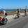 Navy Sweeps Men and Women Team Golds at 2021 Armed Forces Triathlon A group of cyclists are competing in what appears to be a triathlon race along a coastal road. Five riders are visible in a pack, wearing triathlon gear including helmets and form-fitting suits. The setting features a bay or estuary with marshland vegetation and an industrial skyline visible in the background.