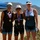 Navy Sweeps Men and Women Team Golds at 2021 Armed Forces Triathlon Three female athletes pose together on a sandy beach after completing a triathlon or similar endurance race. Each woman is wearing a medal around her neck and athletic race gear representing different U.S. military branches, including the U.S. Air Force and U.S. Marines. The setting appears to be a coastal beach location with mountains and blue water visible in the background.