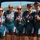 Navy Sweeps Men and Women Team Golds at 2021 Armed Forces Triathlon Five women wearing matching Navy triathlon uniforms pose together outdoors, each wearing medals on red, white, and blue ribbons. The uniforms are teal with gold lettering reading 'NAVY' and feature American flag sleeve accents, branded by Pearl Izumi. The setting appears to be an outdoor athletic competition venue near a body of water with hills in the background.