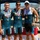 Navy Sweeps Men and Women Team Golds at 2021 Armed Forces Triathlon Three male athletes pose together under a canopy tent at what appears to be a military triathlon competition. Two athletes are wearing Navy team tri-suits and one is wearing a U.S. Air Force tri-suit, all three displaying medals around their necks. The setting appears to be an outdoor venue near a body of water with mountains visible in the background.