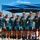 Navy Sweeps Men and Women Team Golds at 2021 Armed Forces Triathlon A group of nine male athletes wearing matching teal 'NAVY' cycling/triathlon uniforms pose together under a blue canopy tent. Most members are wearing medals around their necks, suggesting they recently competed in and placed at a sporting event. The setting appears to be an outdoor competition venue with hills and clear blue sky visible in the background.