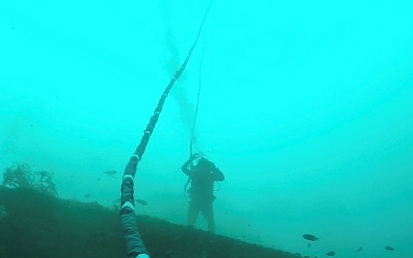 An underwater photograph showing a scuba diver in the background near what appears to be a sloped seafloor or submerged structure. A large cable or hose runs diagonally across the foreground from bottom-right to upper-center of the frame. The water has low visibility with a characteristic blue-green tint typical of murky underwater environments.