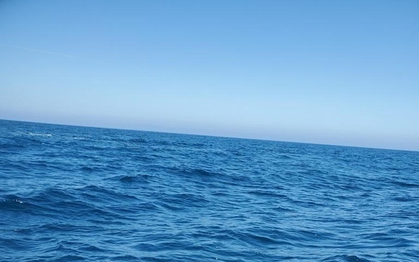 Two individuals are aboard a black inflatable rigid hull boat (RHIB) on open ocean water, handling a red underwater remotely operated vehicle (ROV). The person on the right is wearing a navy blue long-sleeve shirt with partial text visible, suggesting a military or law enforcement affiliation. They appear to be preparing to deploy or recovering the ROV equipment.