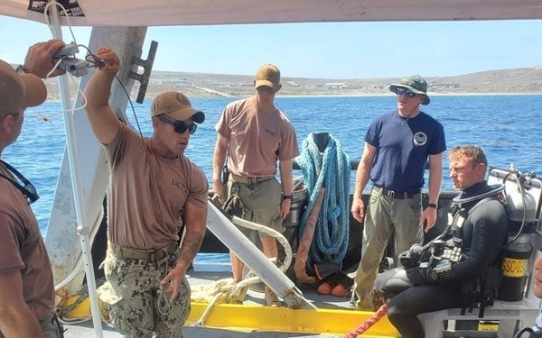 A group of military personnel are gathered on the deck of a vessel, appearing to conduct a pre-dive briefing or equipment check. At least two individuals are wearing full wetsuits with diving equipment (BARE brand), while others are in military utility uniforms and civilian-style tactical clothing. The setting appears to be a coastal or island environment with calm blue water visible in the background.