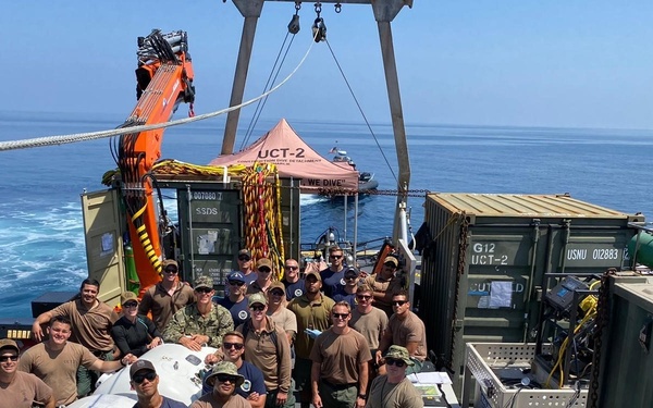 A group of approximately 20 military personnel pose for a group photo on the deck of a naval vessel at sea. The unit is identified as UCT-2 (Underwater Construction Team 2) based on visible markings on equipment containers and a canopy tent. The deck is equipped with diving and construction equipment, suggesting an active underwater construction or salvage operation.