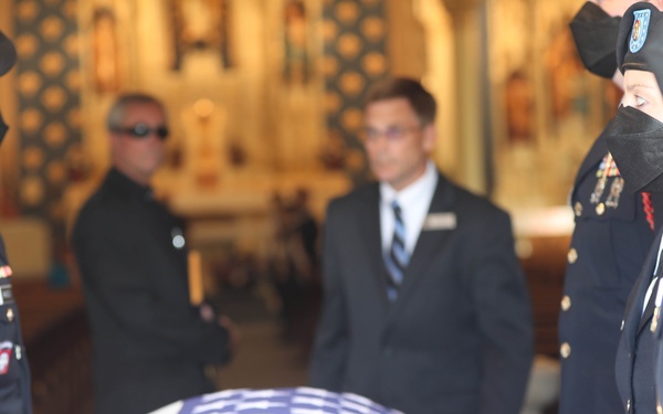 A military funeral ceremony is taking place inside what appears to be a church or chapel. A flag-draped casket covered with an American flag is being attended by uniformed military personnel and civilians. The solemn occasion features soldiers in dress uniforms with visible rank insignia and service ribbons.