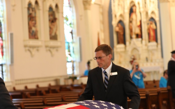A military funeral service is taking place inside a Catholic church, with a flag-draped casket being guided down the center aisle by two men in dark suits. The American flag is fully draped over the casket in the traditional military honors style. The church interior features stained glass windows, wooden pews, and religious iconography typical of a Catholic sanctuary.