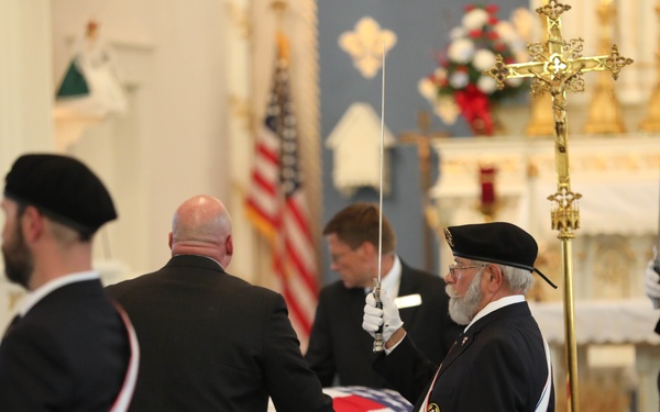 A formal military or fraternal honor guard ceremony is taking place inside a Catholic church. Members wearing black berets, dark suits, white gloves, and red-white-blue sashes are holding raised swords over a flag-draped casket. The setting includes church pews, an American flag, a large gold crucifix, and a decorated altar in the background.