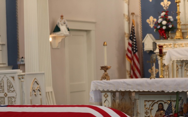 A flag-draped coffin is positioned in the nave of a Catholic church, indicating a military funeral service. The church features ornate white and gold altar furnishings, religious statuary, and an American flag displayed near the sanctuary. The setting suggests a formal funeral Mass honoring a deceased military veteran.