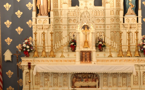 A military funeral service is taking place inside a Catholic church, with a flag-draped casket positioned before the altar rail. The ornate white and gold altar features candles, floral arrangements, and religious statuary against a blue wall decorated with gold fleur-de-lis motifs. An American flag and what appears to be a papal or Vatican flag are displayed on either side of the sanctuary.