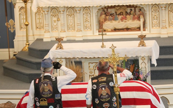 A military funeral service is taking place inside a Catholic church, with a flag-draped casket positioned before the altar. Two men wearing black leather vests adorned with patches — including visible 'Vietnam Veteran' insignia — are rendering a salute to the deceased. Mourners are seated in the church pews observing the solemn ceremony.