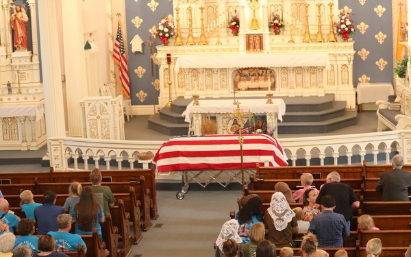 A military funeral service is being held inside a Catholic church, with a flag-draped casket positioned at the front near the altar. Many attendees are wearing matching bright blue t-shirts that read 'Home At Last' on the back, suggesting an organized group honoring the deceased. The ornate white and blue church interior features religious statues, a prominent altar, and American flags flanking the sanctuary.