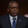 SECDEF, CJCS, CENTCOM Senate Armed Services Testimony A man in formal business attire is seated and speaking, likely at an official hearing or formal proceeding. He is wearing a dark navy pinstripe suit with a blue tie and a small American flag lapel pin. The setting appears to be an official government or institutional venue based on the formal backdrop and microphone placement.