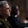 SECDEF, CJCS, CENTCOM Senate Armed Services Testimony The image shows two individuals seated at what appears to be a formal hearing or testimony setting. The foreground figure is wearing a U.S. Army military uniform with an 'Airborne' patch and 101st Airborne Division insignia visible. A second individual in a dark suit is seated in the background, slightly out of focus.
