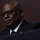SECDEF, CJCS, CENTCOM Senate Armed Services Testimony A man in formal business attire is photographed in a dramatic, low-key lighting setting against a dark background. He is wearing a dark suit, white dress shirt, blue tie, and dark-framed glasses. A small American flag pin is visible on his lapel.