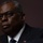 SECDEF, CJCS, CENTCOM Senate Armed Services Testimony A close-up portrait of a man wearing dark-framed glasses, a dark suit jacket, white dress shirt, and a blue tie with a small American flag pin visible on the lapel. The image is dramatically lit with a dark background, giving it a formal and professional appearance. The subject appears to be speaking or gesturing, with one hand partially visible in the lower left.