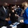 SECDEF, CJCS, CENTCOM Senate Armed Services Testimony A formal meeting or hearing is taking place in what appears to be a government or congressional setting. Several individuals wearing black face masks are engaged in conversation near a table with a nameplate visible reading 'Austin'. The setting includes military personnel in uniform and civilians in formal attire.