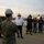 Firefighters Tour Submarine A briefing or coordination meeting is taking place at a naval shipyard or waterfront facility between U.S. Navy personnel and civilian firefighters. A Navy sailor in camouflage uniform and white hard hat is addressing a group that includes a fire department officer in white uniform, firefighters in turnout gear with numbered helmets, and additional personnel. The setting appears to be a pier or dock area during early morning or evening hours.