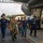 Firefighters Tour Submarine A group of personnel wearing face masks and hard hats walk through what appears to be a naval vessel's hangar bay or dry dock facility. A uniformed U.S. Navy sailor in camouflage utilities appears to be leading or briefing the group, which includes individuals wearing firefighter helmets. The setting suggests a tour, inspection, or training exercise aboard or near a large naval vessel.
