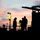 Firefighters Tour Submarine A silhouetted scene of three workers gathered on what appears to be an industrial platform or construction site at dusk or dawn. The workers are wearing hard hats and appear to be in discussion or reviewing equipment. A single illuminated work light on a pole provides artificial lighting against a dramatic pink and purple sky.