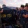 Firefighters Tour Submarine A U.S. Navy sailor in camouflage uniform and white hard hat is briefing a group of civilian firefighters on the deck of a naval vessel. The sailor is holding up a black banner marked 'SSN 773', which identifies the submarine USS Pittsburgh, a Los Angeles-class attack submarine. The scene appears to take place at dusk during what looks like a damage control or emergency response coordination exercise.