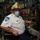 Firefighters Tour Submarine A fire department Deputy Chief, identified by the gold helmet reading 'Deputy Chief Mystic,' is seated inside what appears to be a submarine or naval vessel's control room. He is wearing a white uniform shirt with an EMT patch and a blue surgical face mask. The setting features dense arrays of controls, pipes, gauges, and equipment panels typical of a military vessel interior.