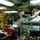 Firefighters Tour Submarine Two military personnel are conducting what appears to be an inspection or equipment check in a shipboard galley or workspace. The individual in the foreground wears Navy digital camouflage (NWU Type III) and a black face mask, holding a gas mask while reaching up toward overhead cables. A second individual in a white uniform and a fire/damage control helmet with a blue face mask observes from behind.