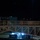Firefighters Tour Submarine A nighttime scene showing multiple uniformed personnel wearing helmets crossing an elevated pedestrian bridge or walkway. Below the bridge, illuminated shipping containers branded 'United Rentals' are visible in what appears to be a parking or staging area. The overall lighting is low, with the primary light sources being the container lights and ambient urban lighting.